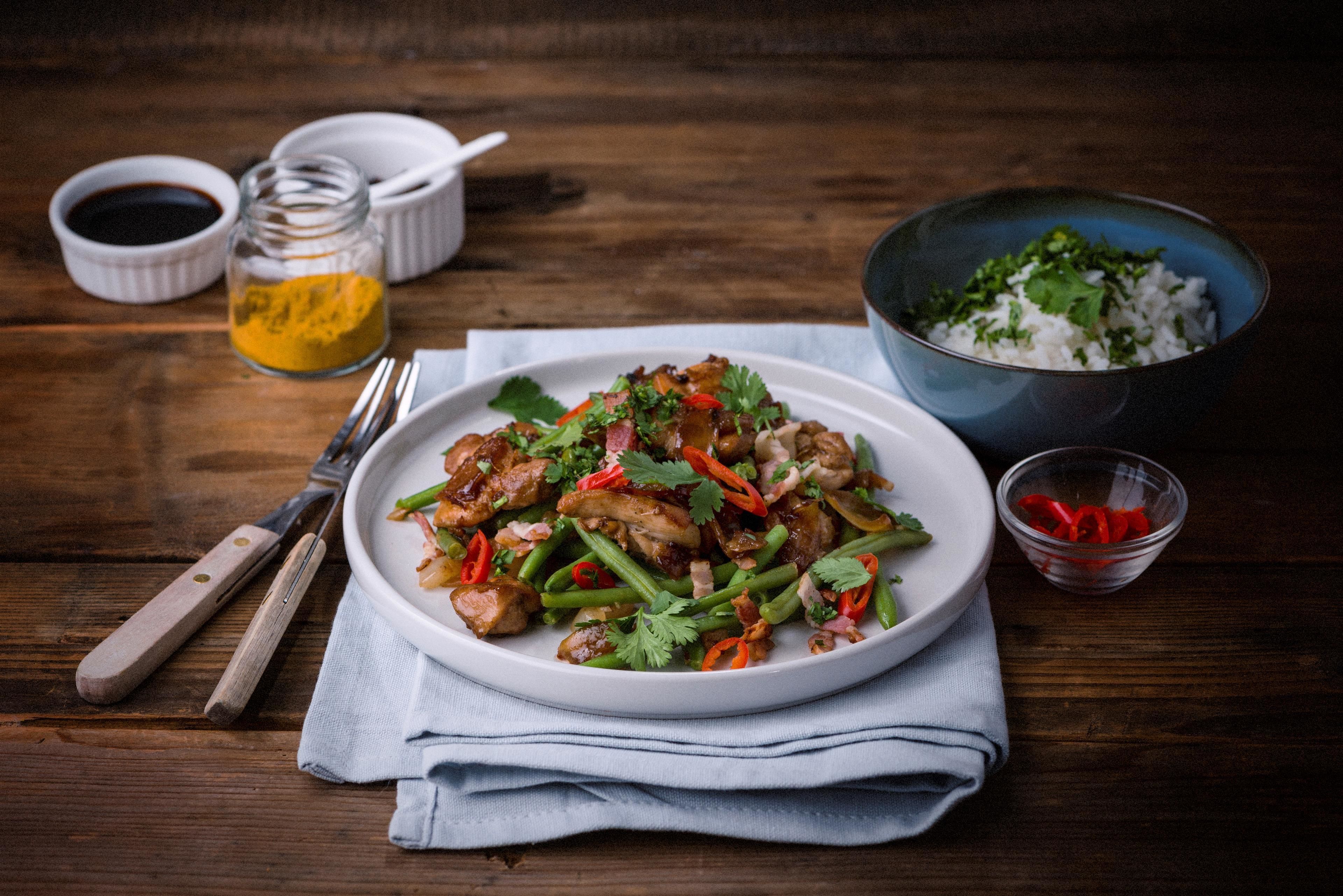 A plate of stir-fried chicken with vegetables on a wooden table, accompanied by bowls of rice and sauces.