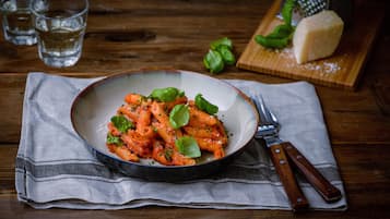 Een kom pasta met basilicum op een houten tafel, samen met een mes en een snijplank met kaas en basilicum.
