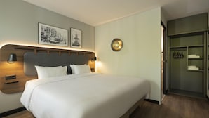 Ein ordentlich eingerichtetes Hotelzimmer mit einem großen Bett, Nachttischlampen, einer Wandleuchte und einem Kleiderschrank mit darin befindlichen Kleidungsstücken.