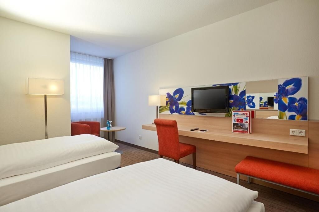 Habitación de hotel con dos camas, un escritorio con televisor de pantalla plana, una silla roja y una mesita con lámpara.