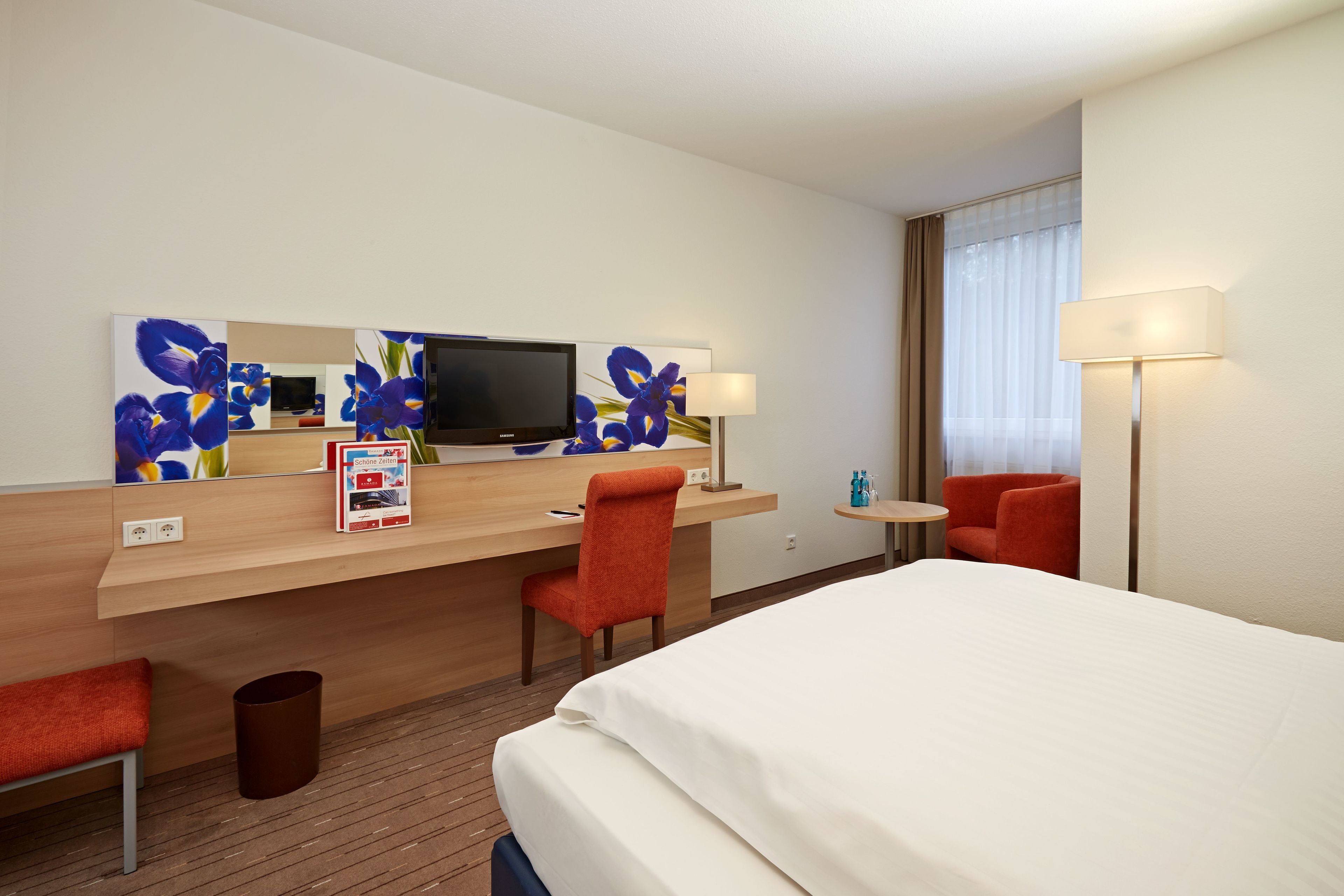 Habitación de hotel con cama, escritorio con televisor, silla, lámpara y ventana con cortinas.