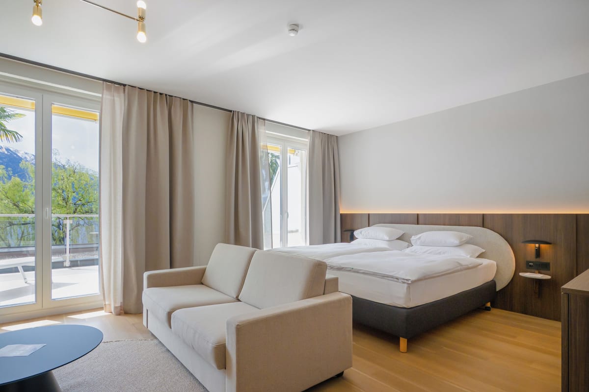 A modern hotel room with a bed, a sofa, a small table, and a view of the outdoors.