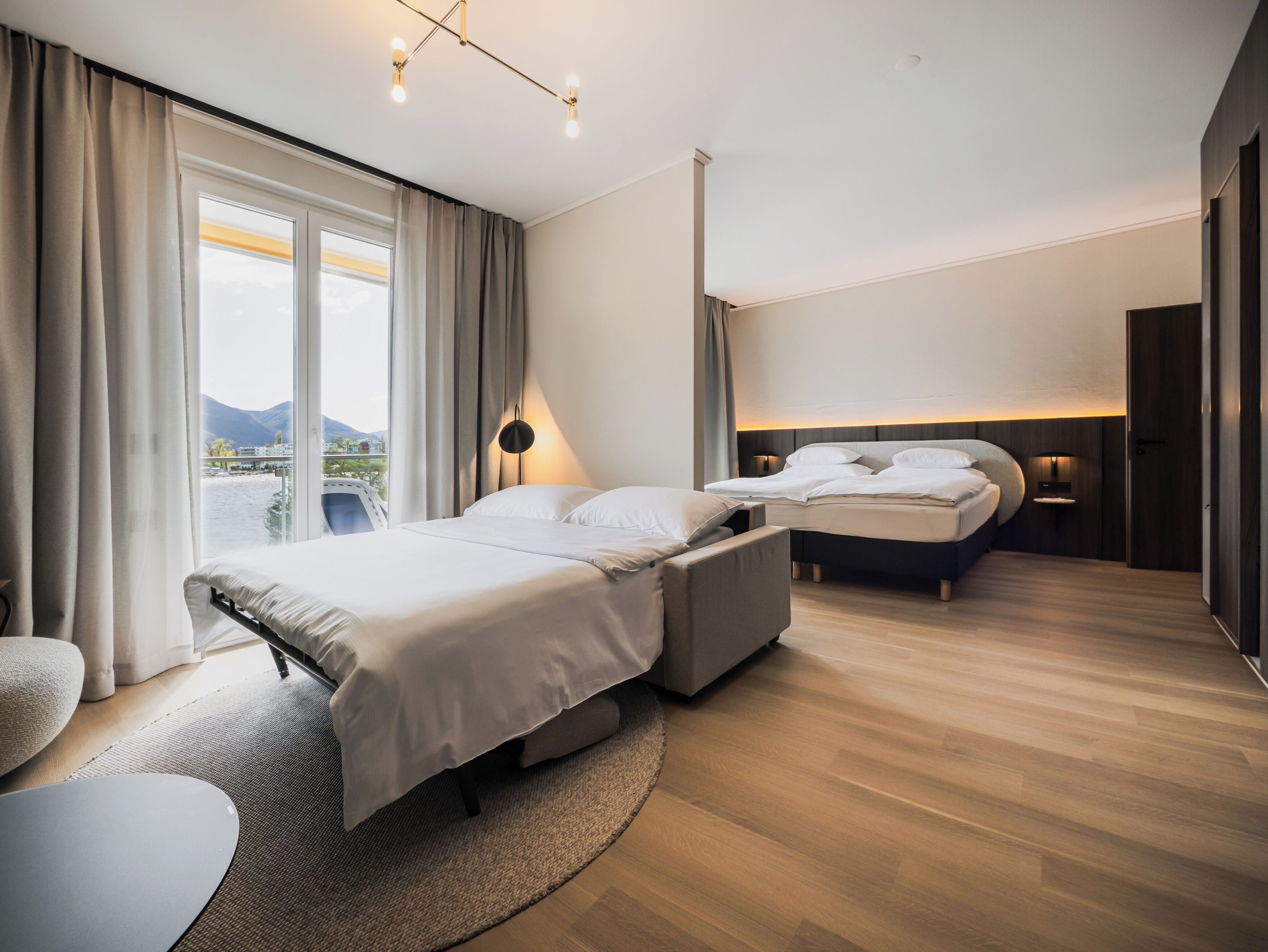 A modern hotel room with a large bed, a smaller bed, a seating area, and a view of the outdoors.