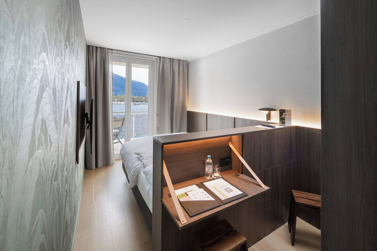 A modern hotel room with a bed, a desk with a lamp, a chair, and a view of a lake and mountains.