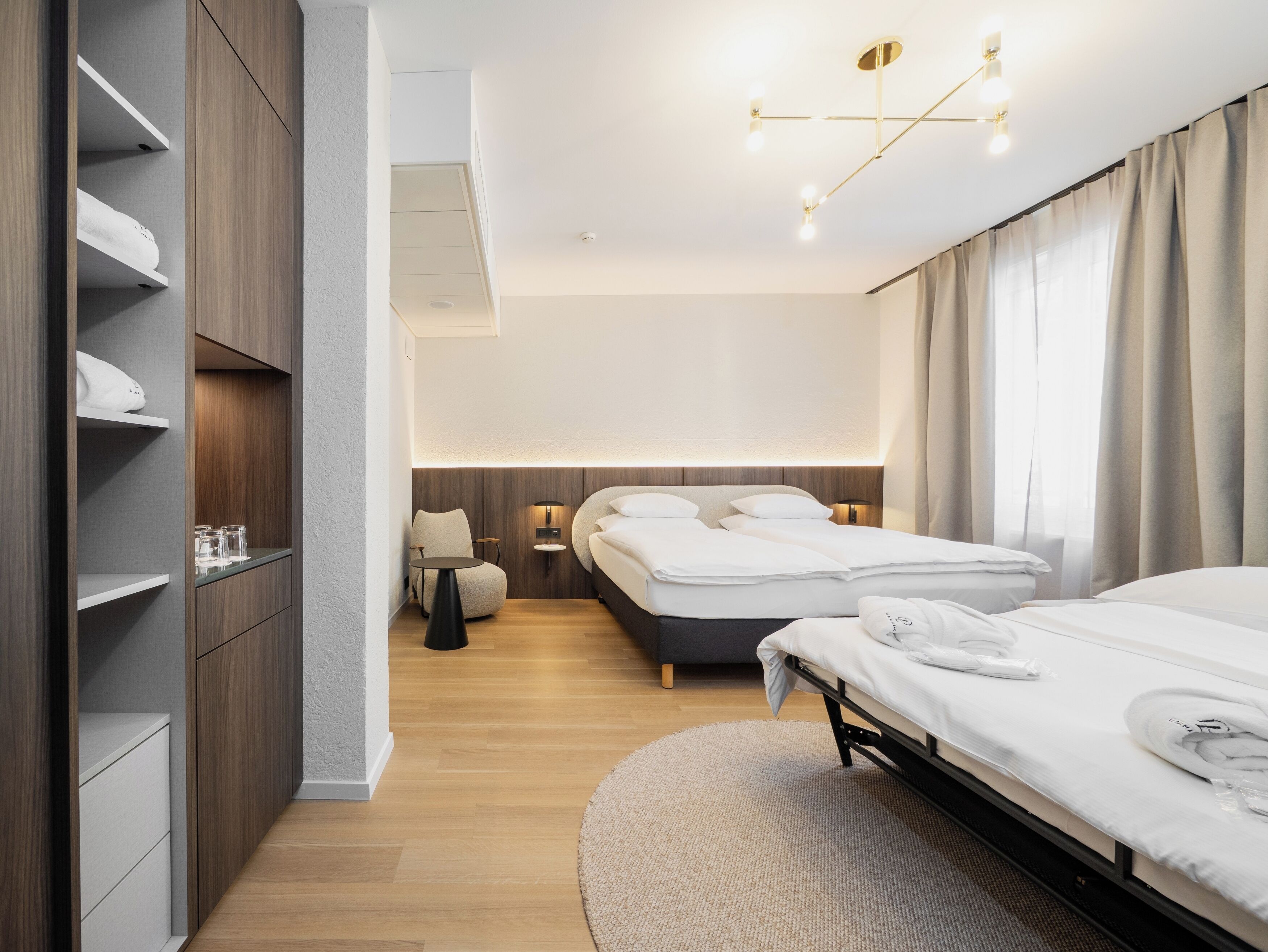 A modern hotel room with a bed, bedside tables, a chair, a wardrobe, and a ceiling light fixture.