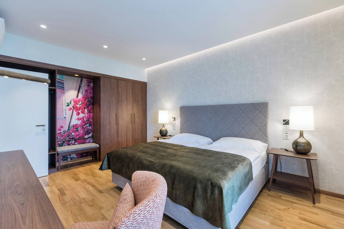 A modern hotel room with a large bed, two bedside tables with lamps, a wooden wardrobe, and a bench with a floral mural.