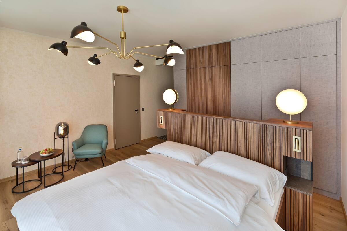 A hotel room with a large bed, a wooden headboard, a round mirror, a blue armchair, and a small table with a fruit bowl and a bottle.