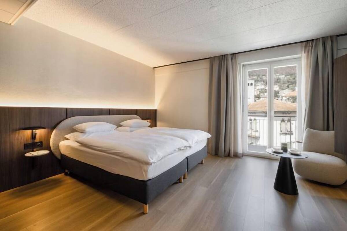 A modern hotel room with a large bed, a bedside table, a small round table, and a view of the city through a large window.