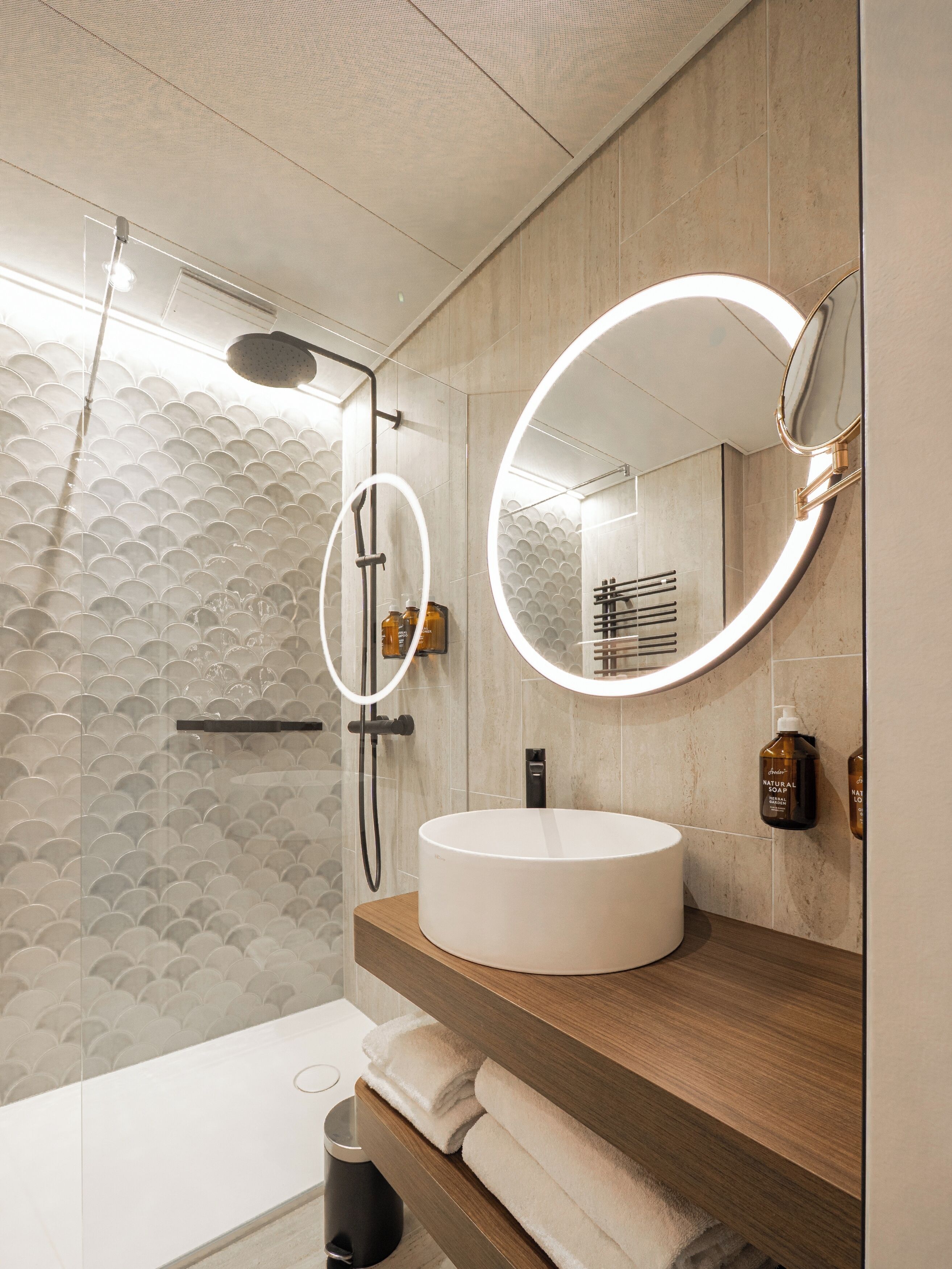 Une salle de bain moderne avec un miroir rond, un lavabo rond et une douche agrémentée d’un mur recouvert de carreaux originaux.