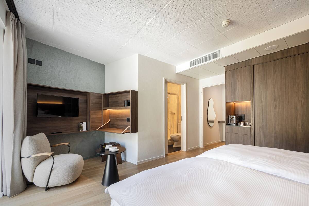 A modern hotel room with a large bed, a seating area with a chair, a TV mounted on the wall, and a wooden wardrobe.