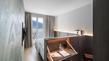 A modern hotel room with a bed, a desk with a lamp, a chair, and a view of a lake and mountains through a large window.