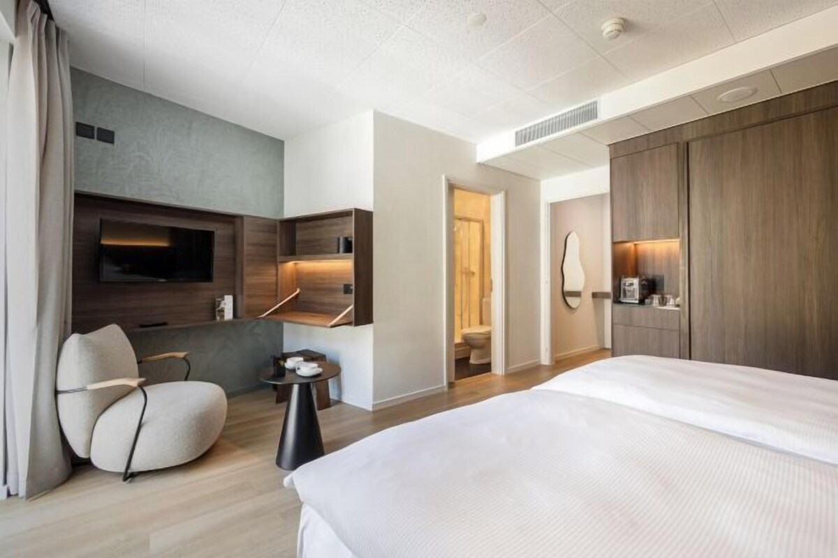 A modern hotel room with a large bed, a seating area with a chair and a small table, a flat-screen TV mounted on the wall, and a wardrobe.