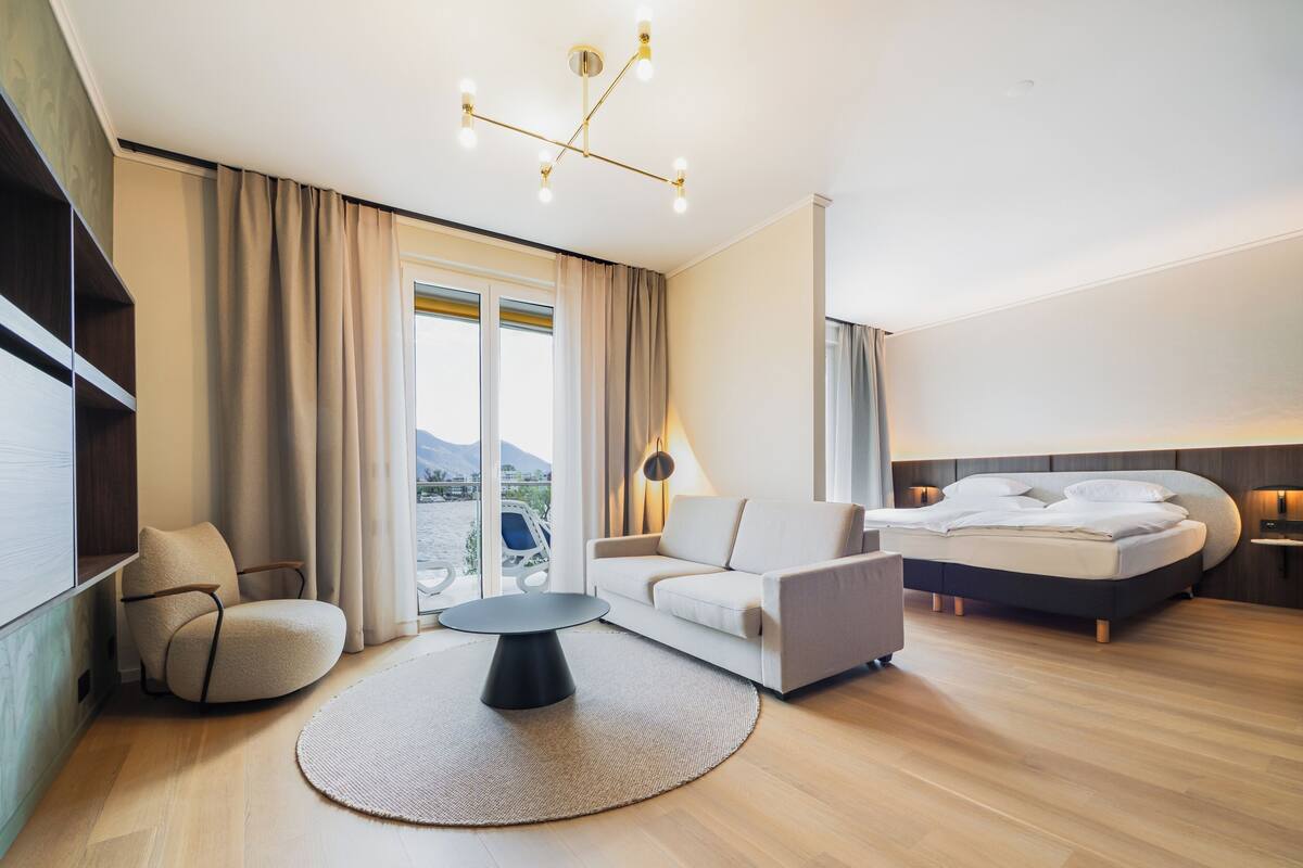 A modern hotel room with a bed, a sofa, a chair, a small table, and a view of a body of water through large windows.