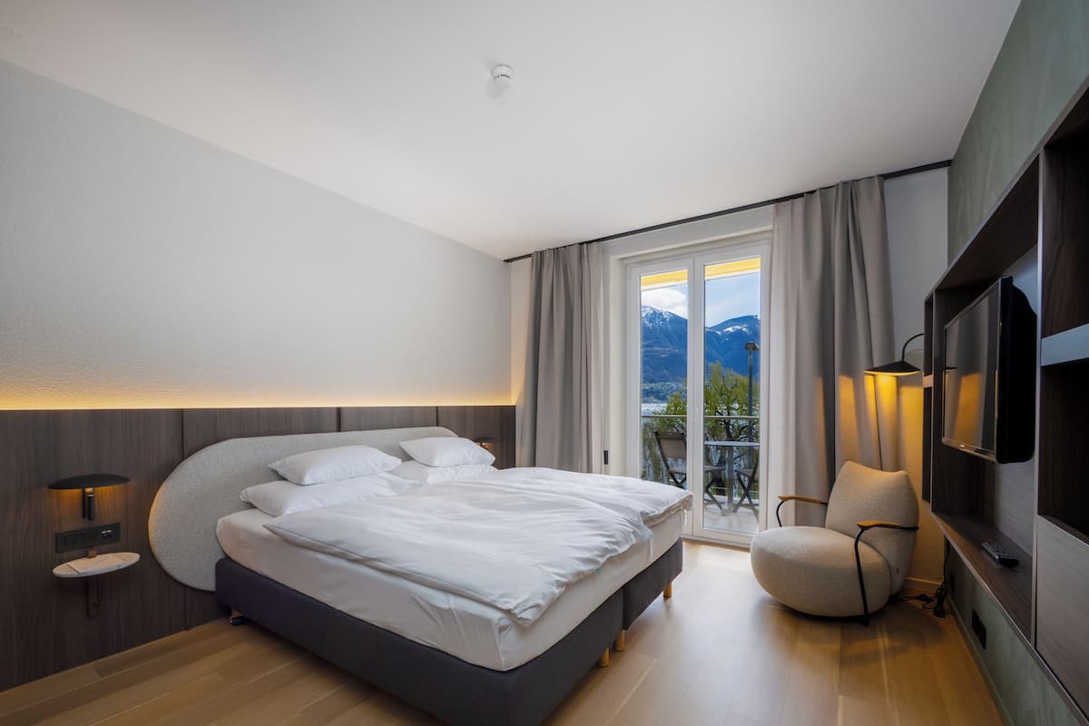 A modern hotel room with a large bed, a TV, a chair, and a balcony with a view of mountains.