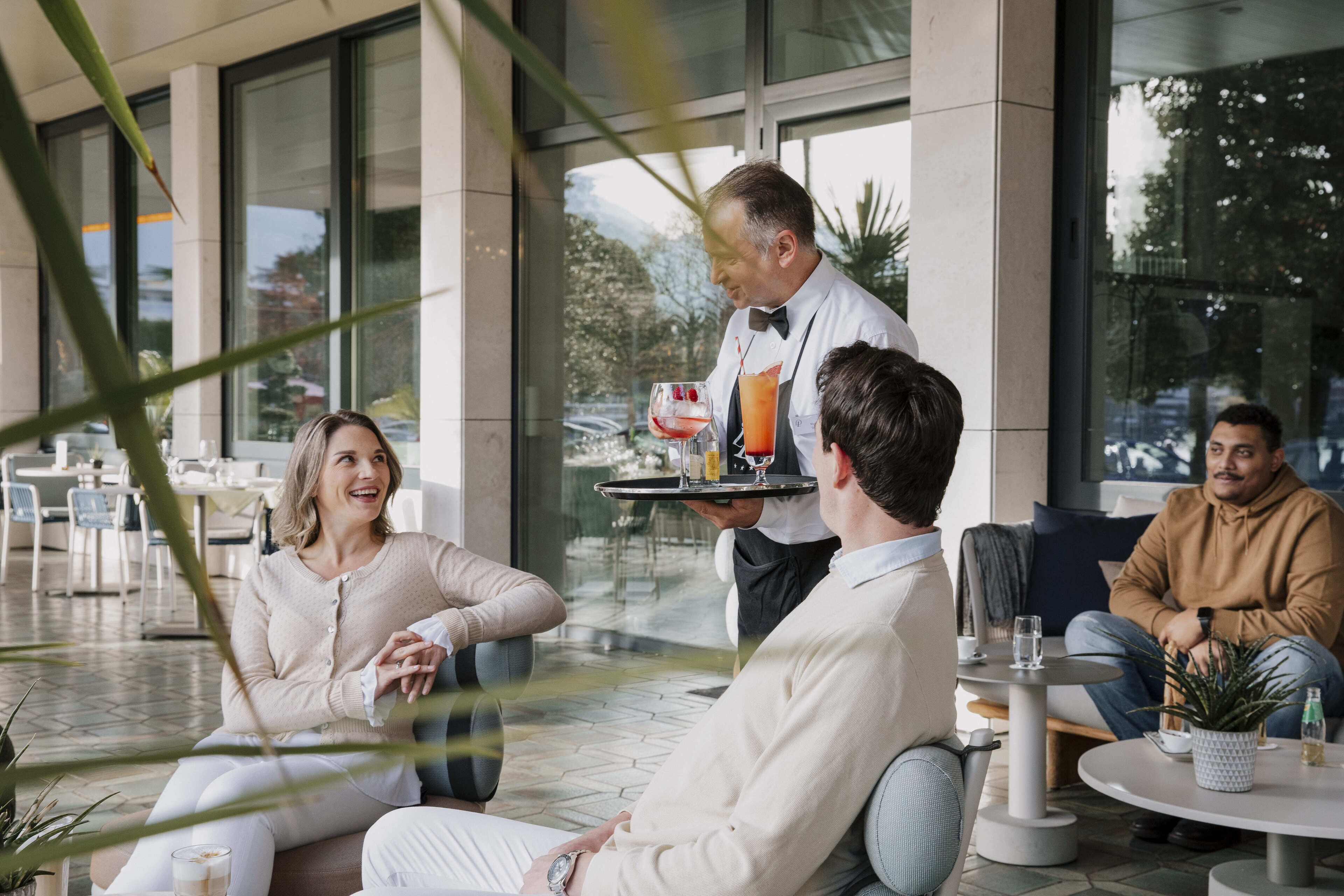 A group of people seated outdoors in a modern setting with large windows, a waiter serving drinks, and a casual dining atmosphere.