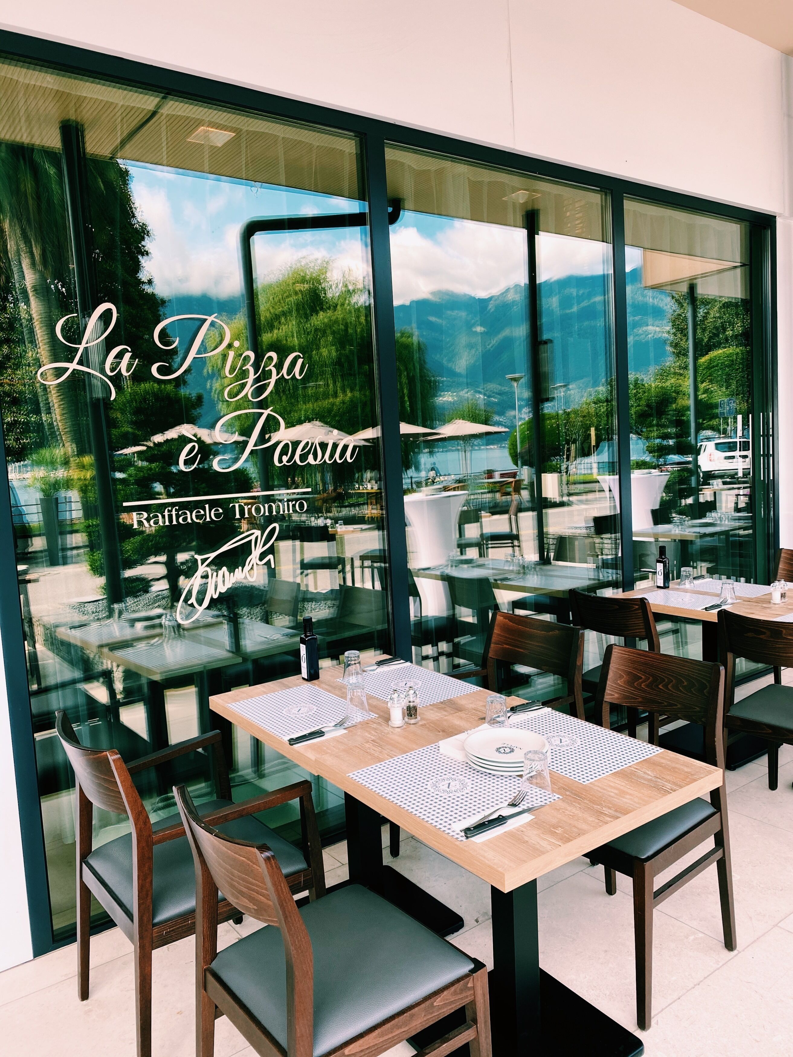 Un espace de restauration en plein air avec des tables et des chaises, un mur de verre qui reflète le ciel et les montagnes, et le nom du restaurant, « La Pizza e Forno », visible sur le verre.