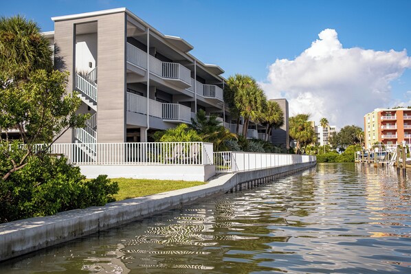 A modern multi-story building with balconies, a concrete waterfront walkway, and surrounding greenery. - The Fold Hotels Indian Shores (Indian Shores)