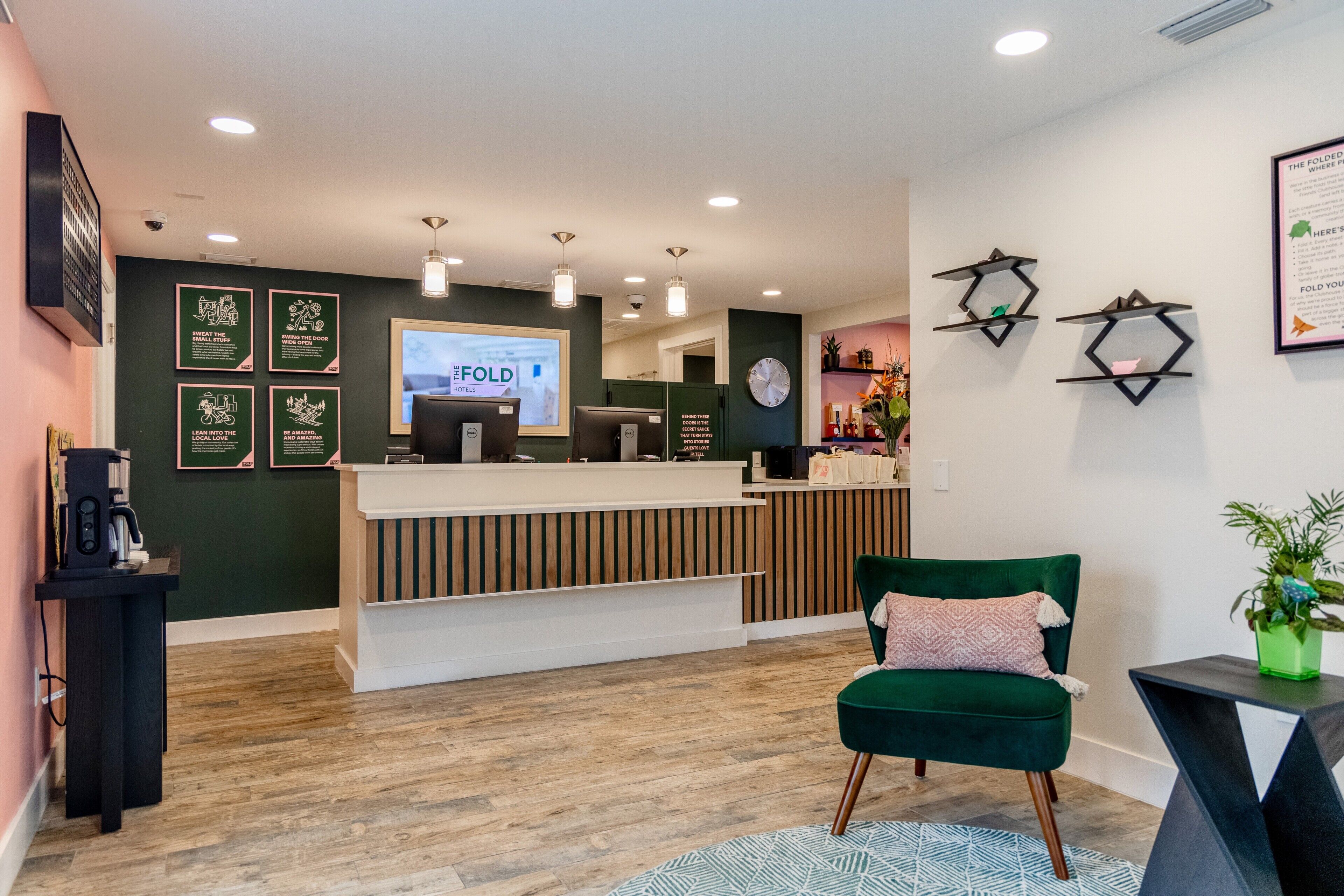 Un hall d’hôtel avec un comptoir d’accueil en bois, une chaise verte, une horloge et un écran mural affichant le nom « THE FOLD HOTEL ».