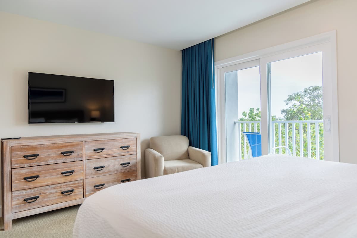 Ein Hotelzimmer mit einem großen Bett, einer Holzkommode, einem an der Wand montierten Fernseher, einem Sessel und einem Balkon mit blauen Vorhängen.