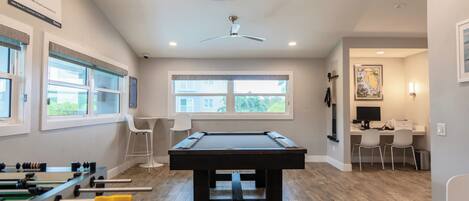 A pool table in a room with a ceiling fan, a foosball table, and a desk with chairs.