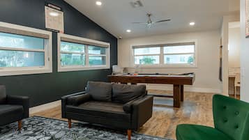 A room with a pool table, a green chair, a black leather sofa, and a ceiling fan.