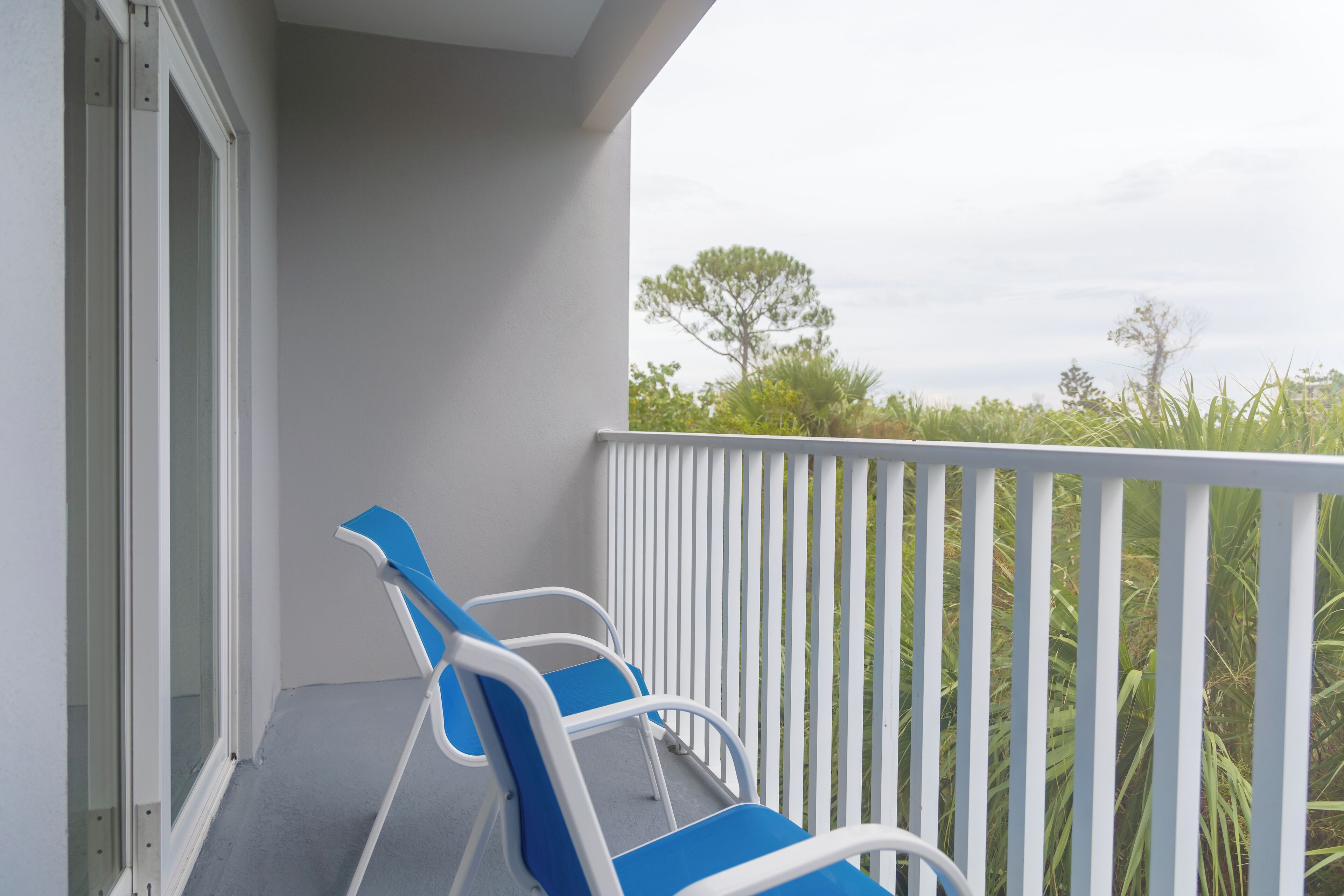 A balcony with two blue and white chairs, a white railing, and a view of greenery and trees.