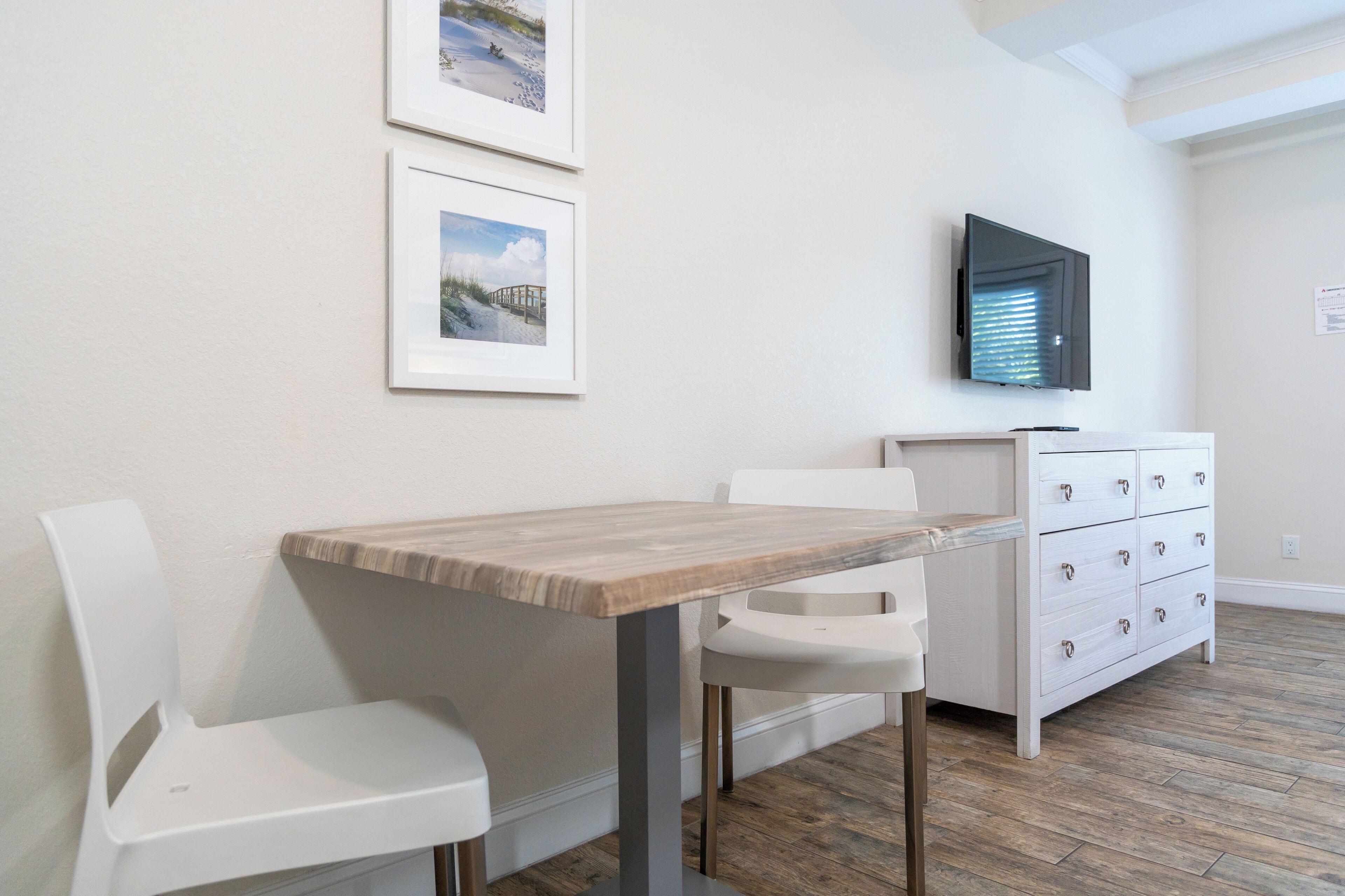 Una habitación moderna con una mesa de madera, sillas blancas, un armario blanco con cajones y dos cuadros en la pared.