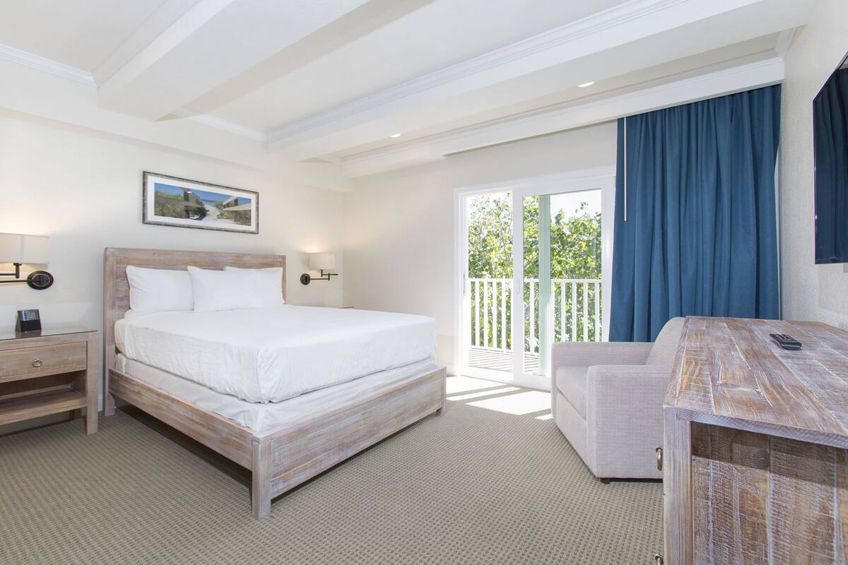 Ein Hotelzimmer mit einem Bett, einem Schreibtisch, einem Sessel, einem Nachttisch, einer Wandleuchte und einem Fenster mit Blick auf Grünflächen.