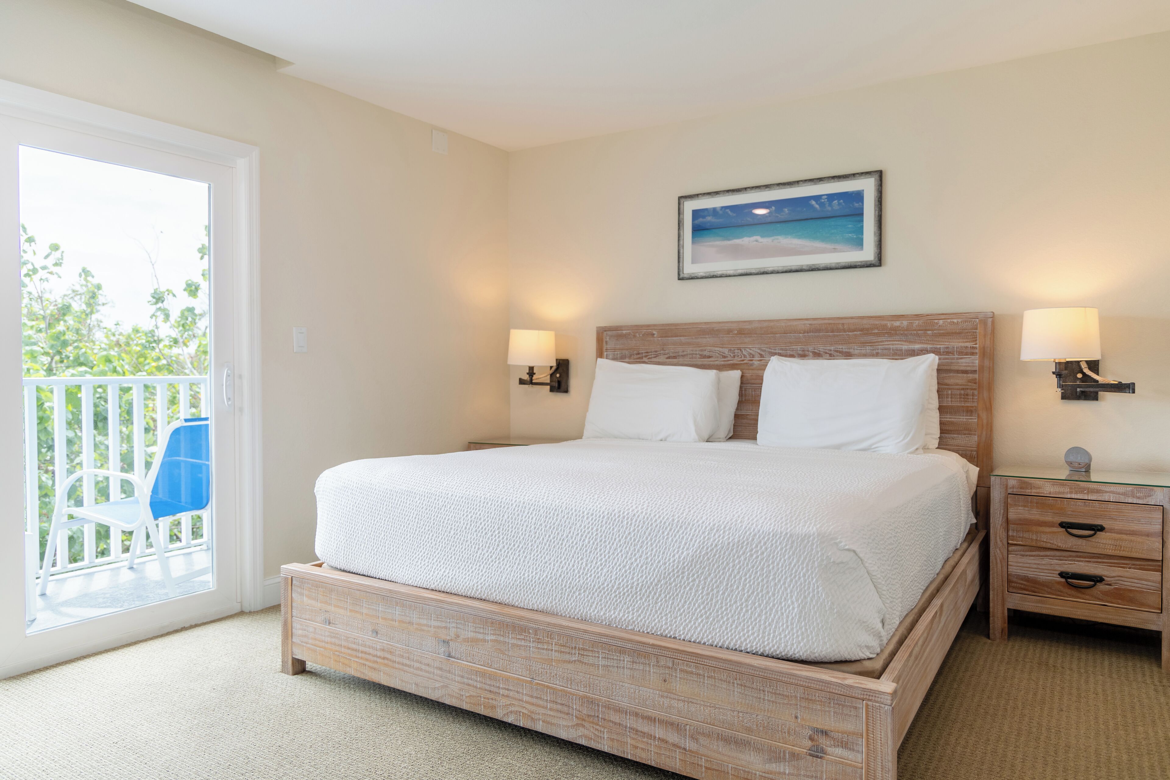 A bedroom with a wooden bed, two bedside tables, a nightstand, a balcony with a chair, and a painting on the wall.