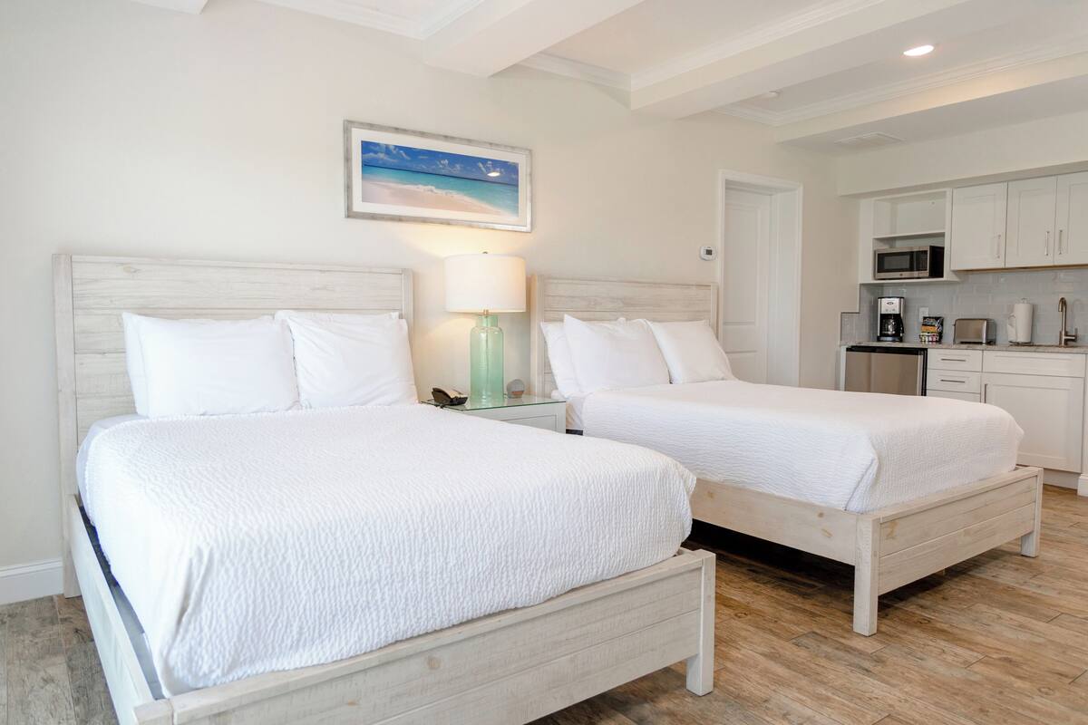 Ein Hotelzimmer mit zwei weißen Betten, einer kleinen Küchenzeile und einem Strandbild an der Wand.
