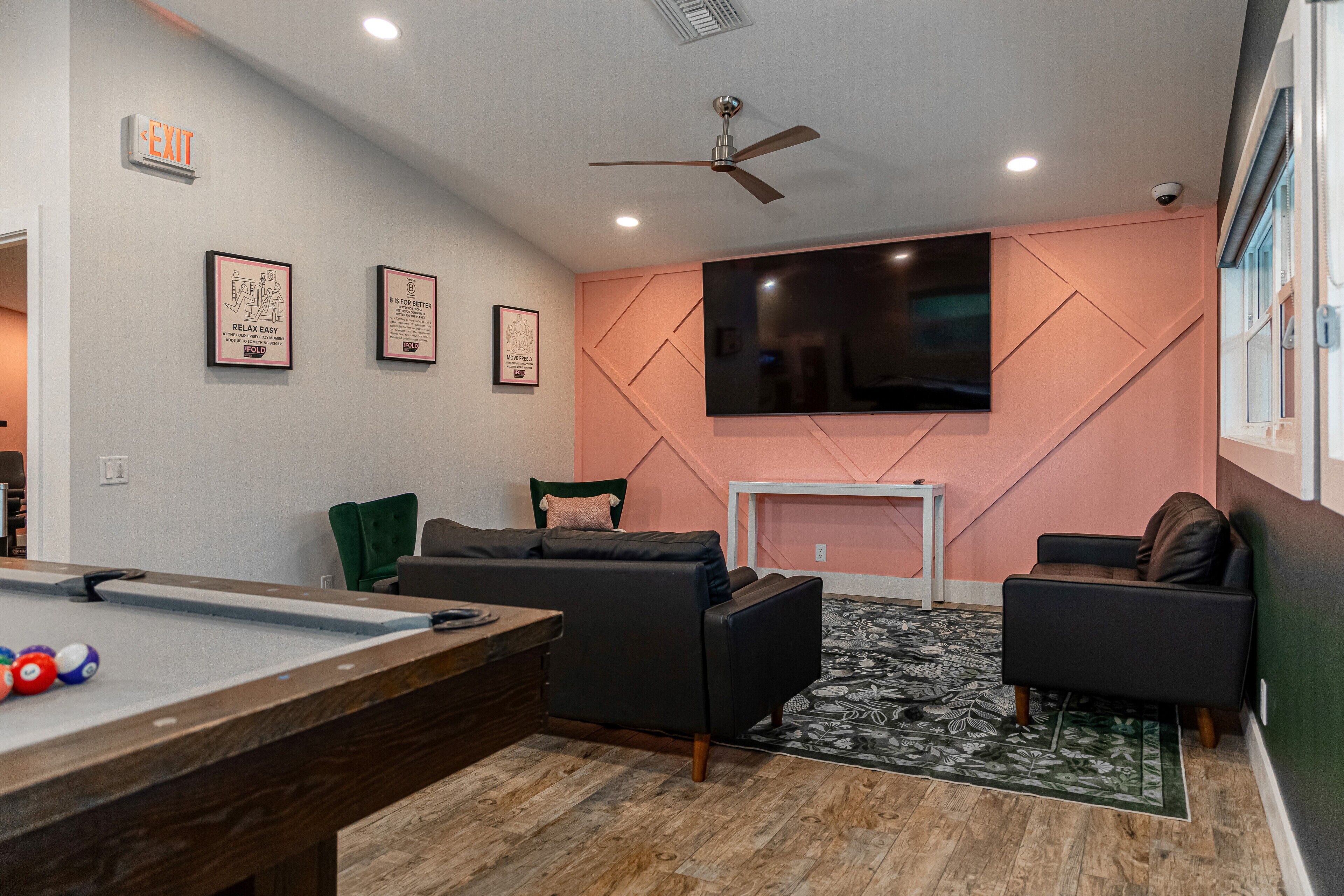 A pool table, two sofas, a television, and a ceiling fan in a room with a pink accent wall.
