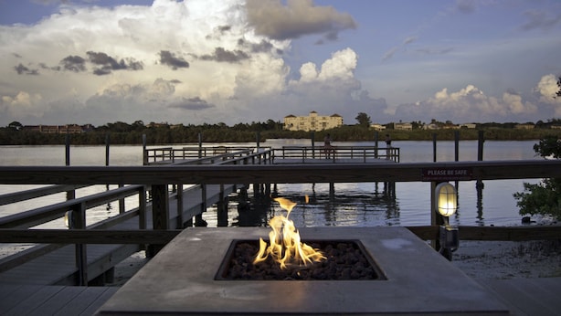 Un muelle de madera con un elemento decorativo de fuego y un cartel que dice 'Por favor, tenga cuidado', con vistas a un lago y un edificio al fondo.