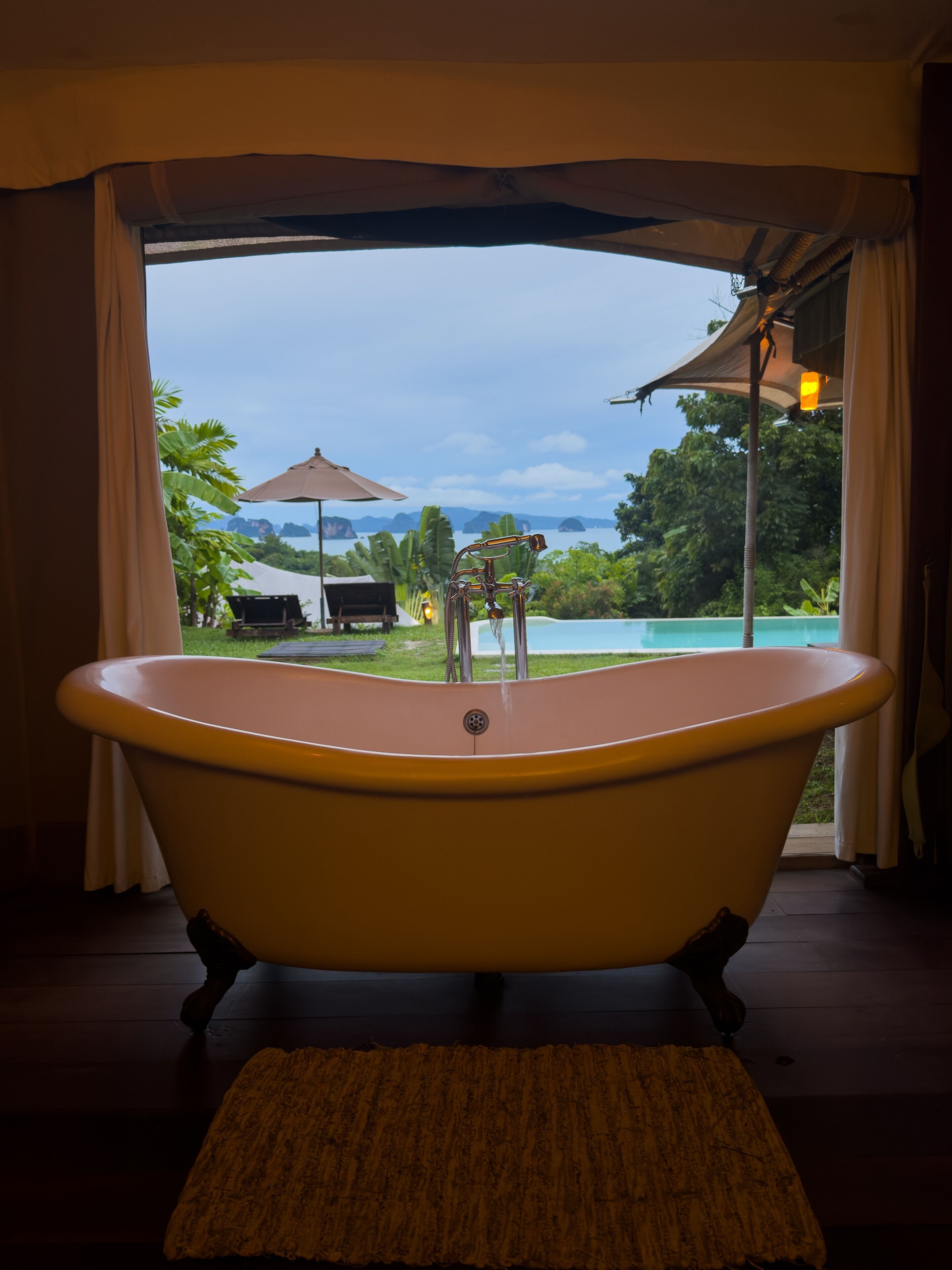 A bathroom with a freestanding bathtub, a view of a pool and tropical landscape through a window.
