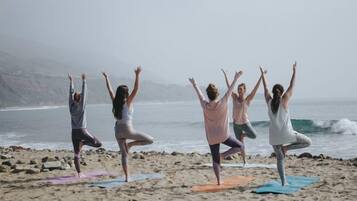 Un grupo de personas practicando yoga sobre esterillas en una playa, con vistas al océano y colinas al fondo.