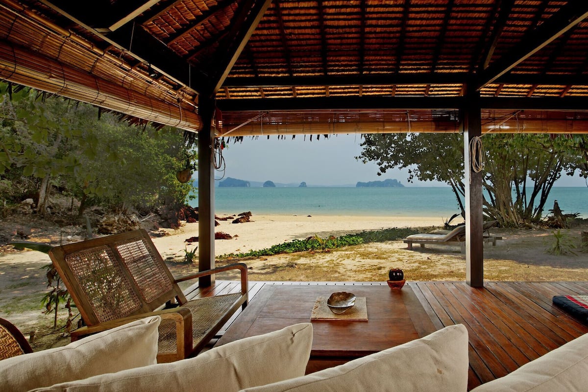 A wooden deck with a table and chairs overlooking a beach with a view of the sea and distant islands.