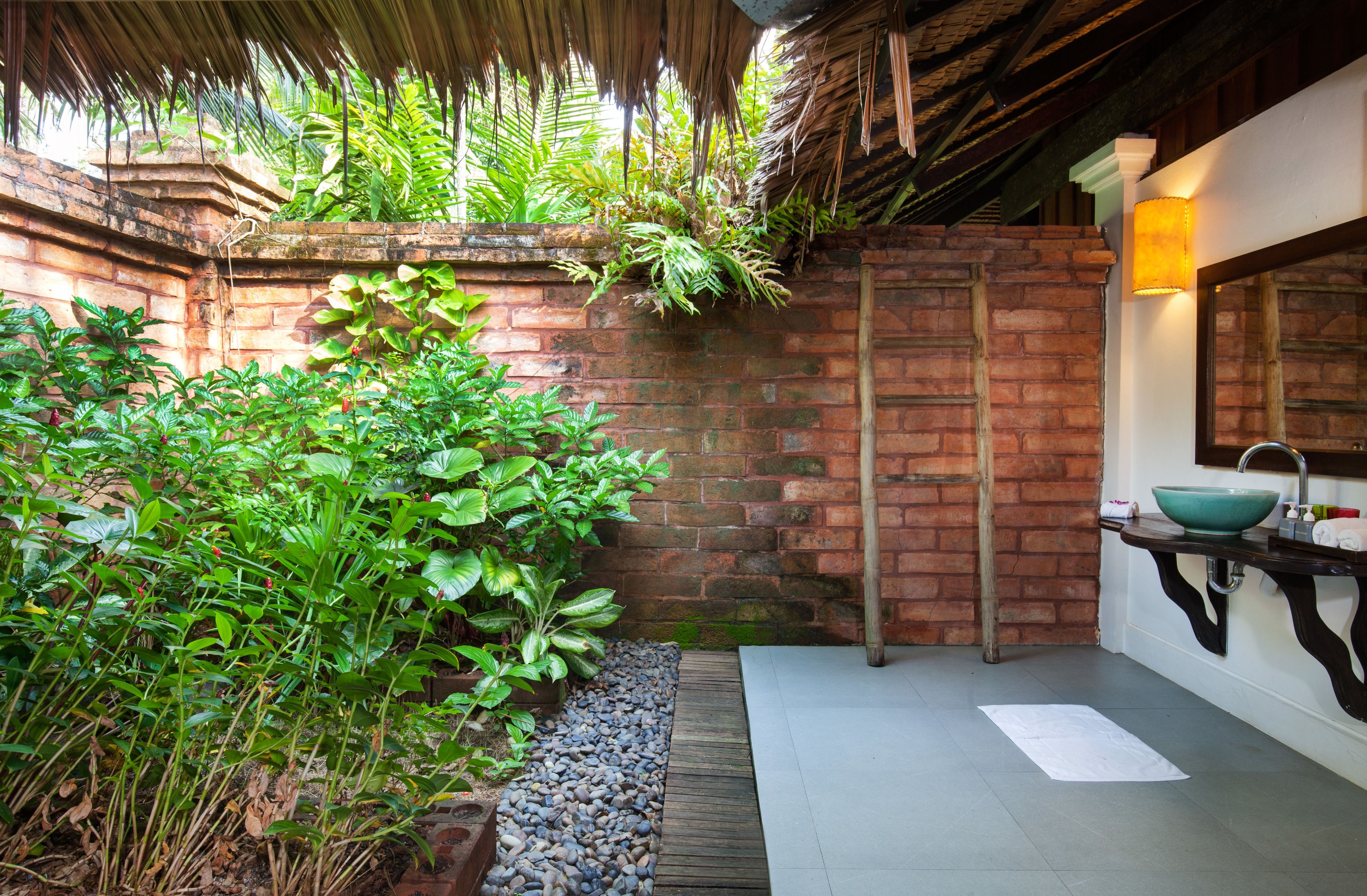 A bathroom with a brick wall, a wooden door, a sink with a green basin, and a thatched roof.