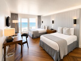 A hotel room with two beds, a desk with a lamp, a chair, a waste bin, and a view of the beach.