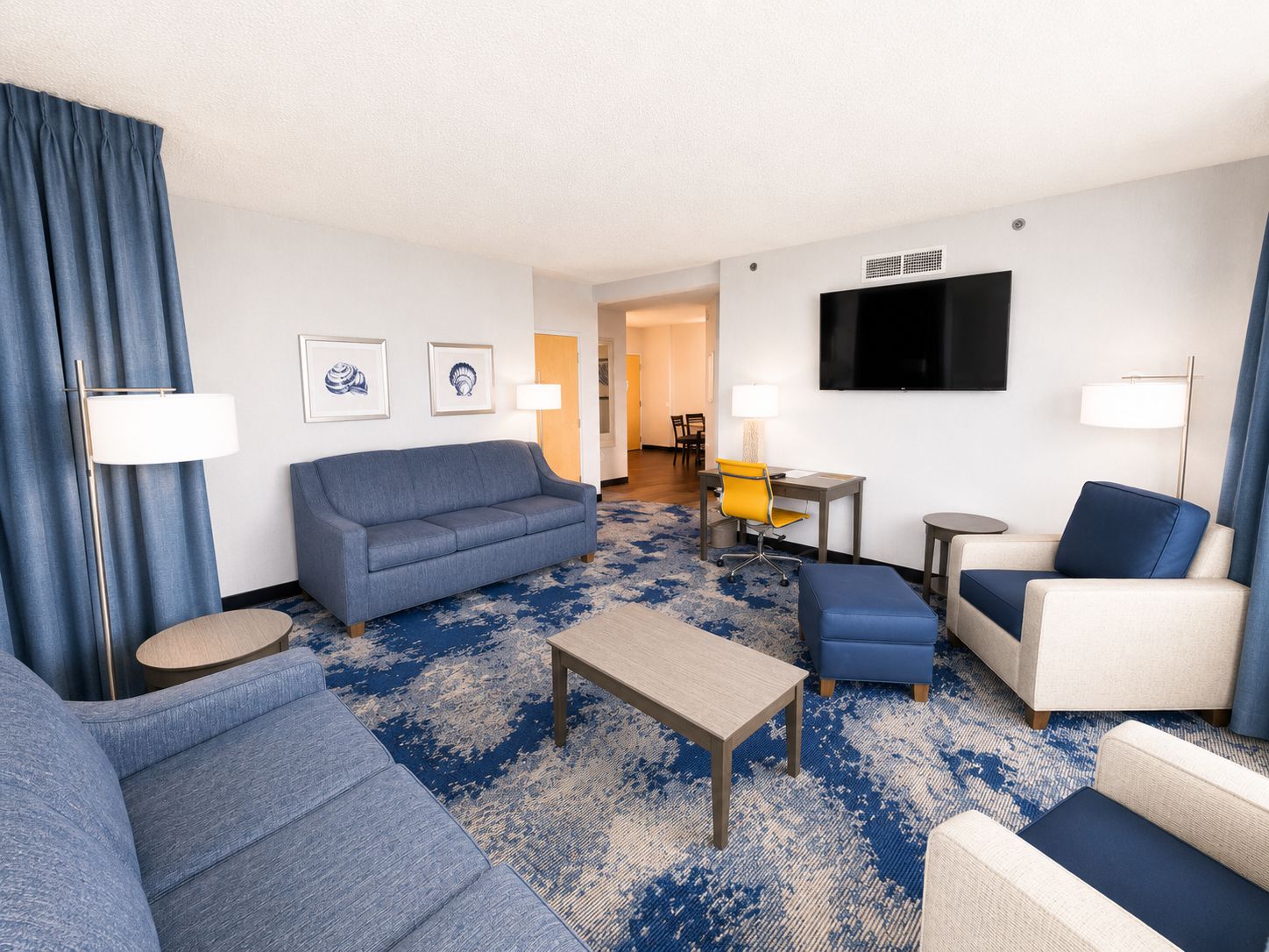 A hotel room with a blue sofa, a small table, a blue armchair, a lamp, and a flat-screen TV on the wall.