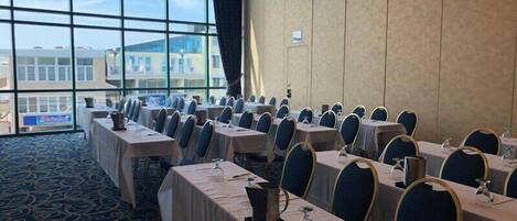 A conference room with rows of tables and chairs, large windows, and a carpeted floor.