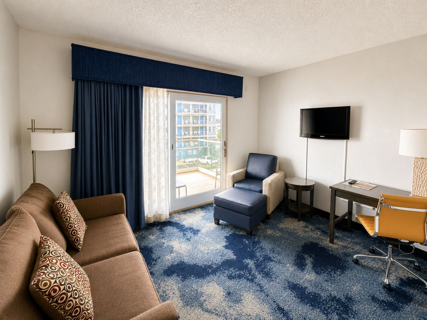 A hotel room with a sofa, armchair, ottoman, desk, TV, and a view of the outside balcony.