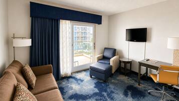A hotel room with a sofa, armchair, ottoman, desk, TV, and a view of the outside balcony.