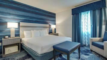 Een hotelkamer met een groot bed, twee nachtkastjes met lampen, een bankje, een sofa en uitzicht buiten door een raam met gordijnen.