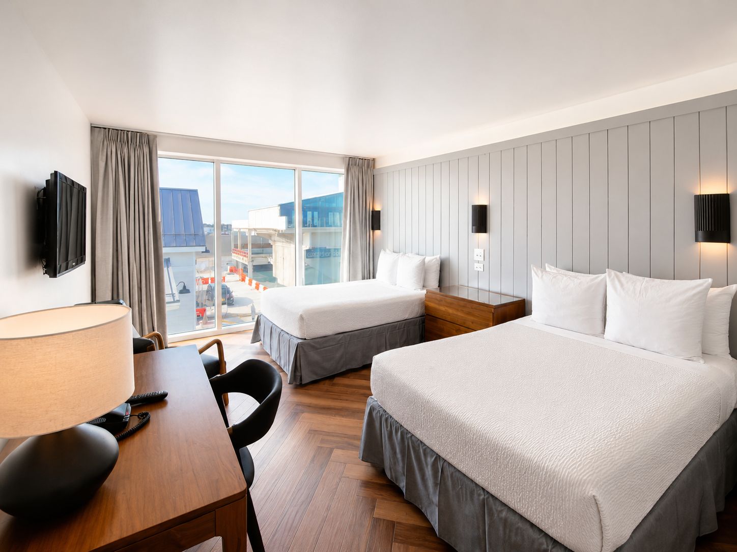 A hotel room with two beds, a desk, a chair, a TV, and a large window with a view of the city.