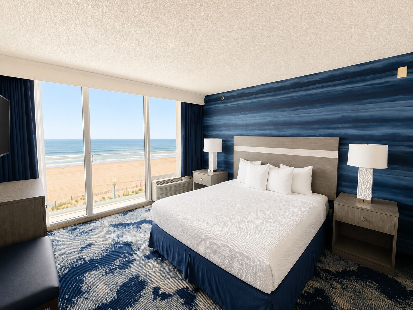 A hotel room with a large bed, two bedside tables with lamps, a view of the beach, and a patterned carpet.