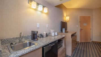 Una cocina de hotel con encimera de granito, pileta de acero inoxidable, cafetera y refrigerador. Hay un microondas, un refrigerador pequeño y un televisor sobre un pedestal. La habitación tiene piso alfombrado y una puerta que da a otra área.