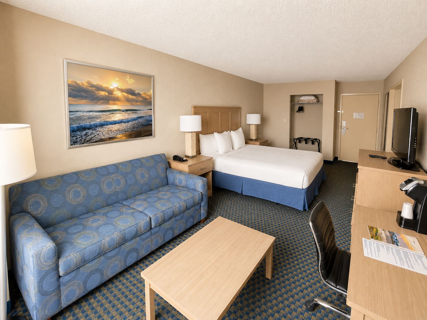 A hotel room with a bed, a sofa, a desk, a television, and a painting of a sunset over the ocean.