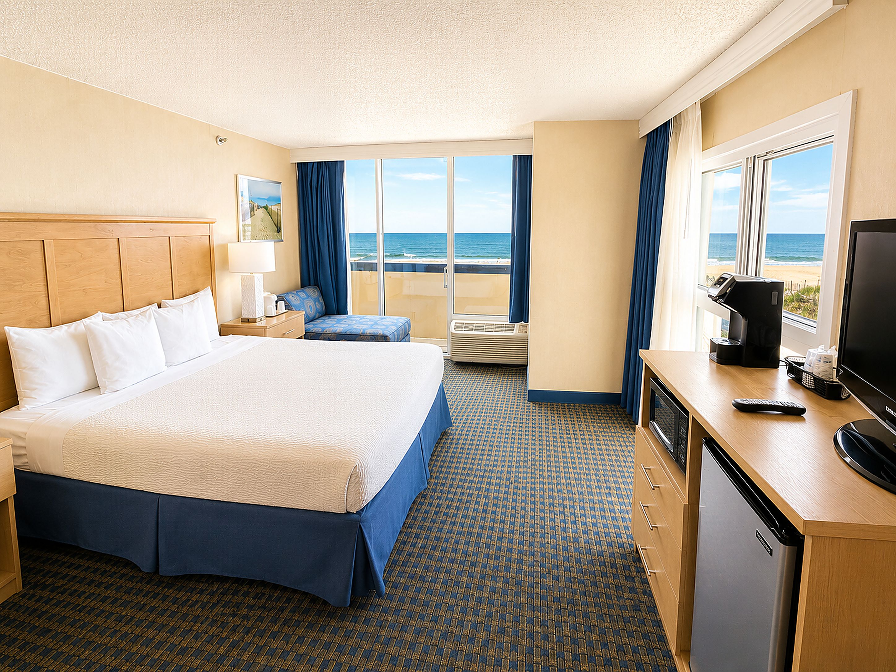 A hotel room with a bed, a desk with a television, a coffee maker, and a view of the ocean.