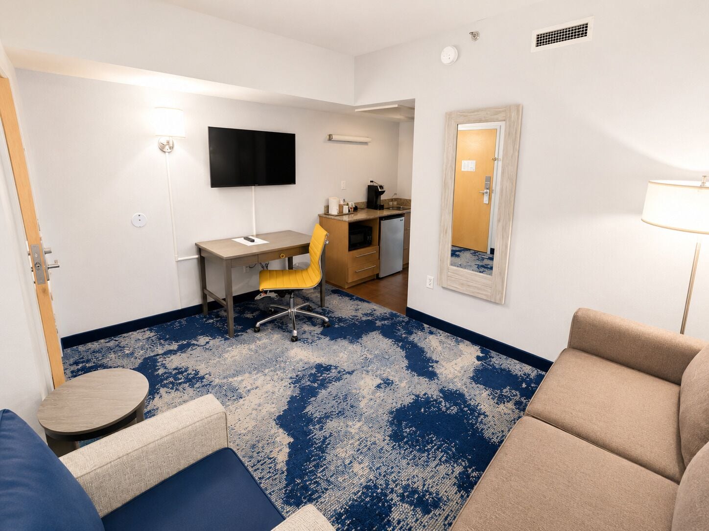 A compact hotel room with a desk, chair, sofa, and a television.