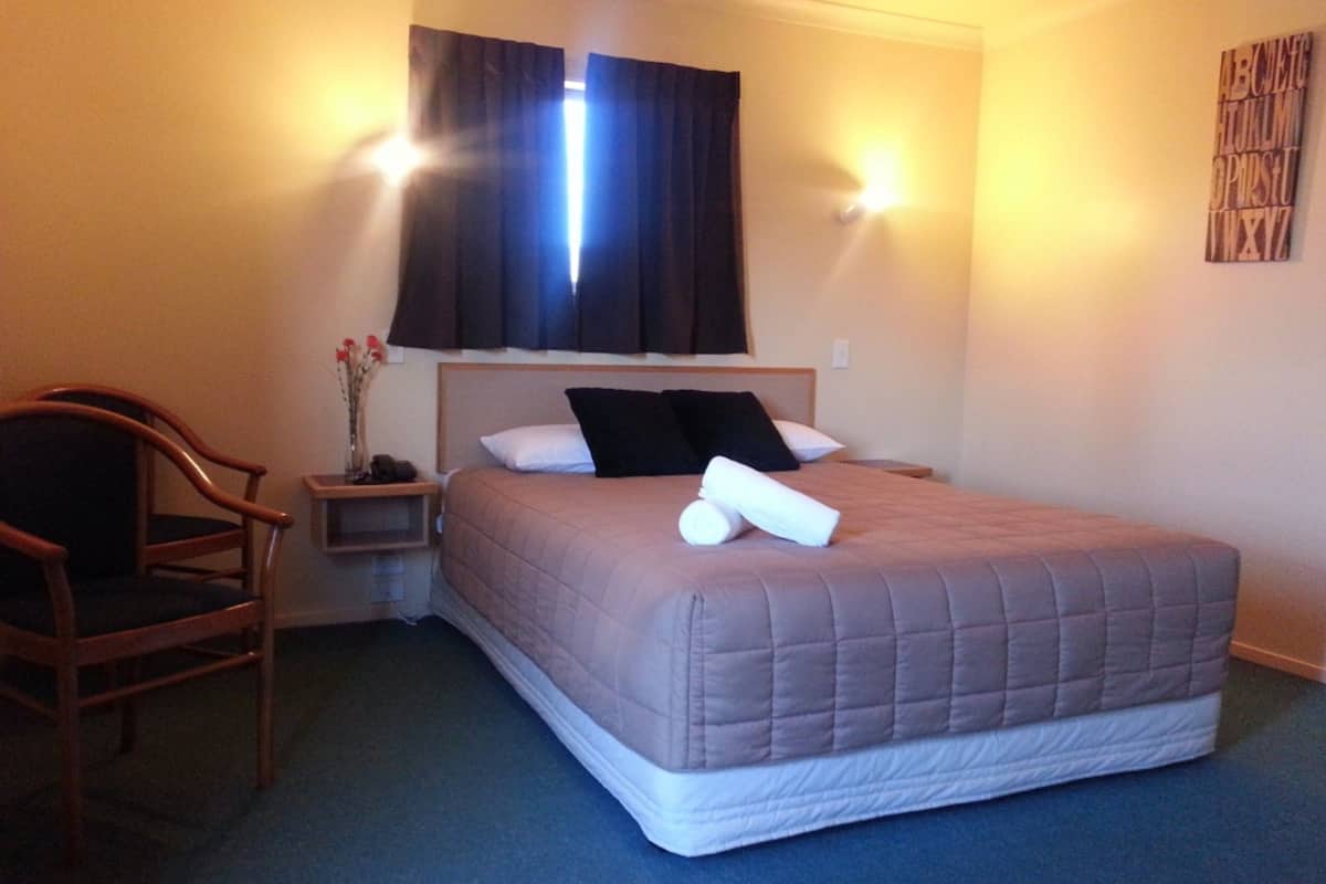 A hotel room with a bed, a chair, a nightstand, and a wall-mounted light fixture.