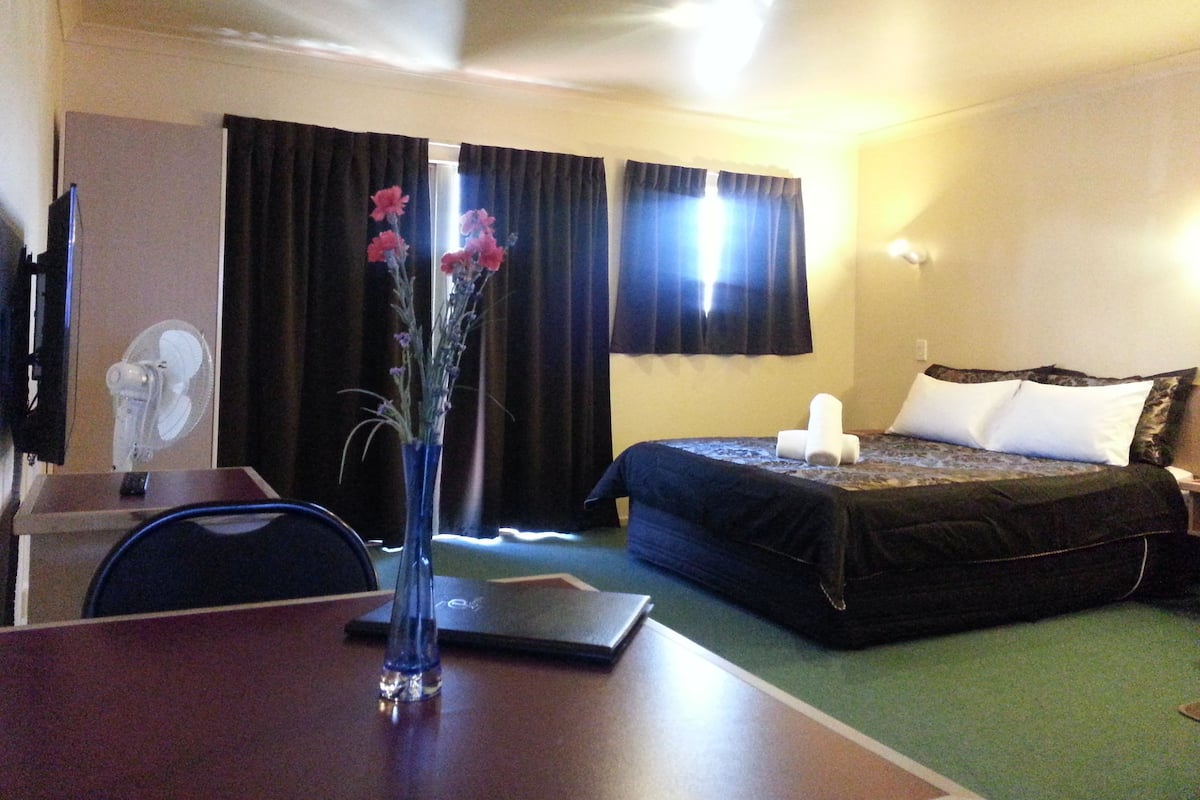 A hotel room with a bed, a desk with a laptop, a vase with flowers, and a television.